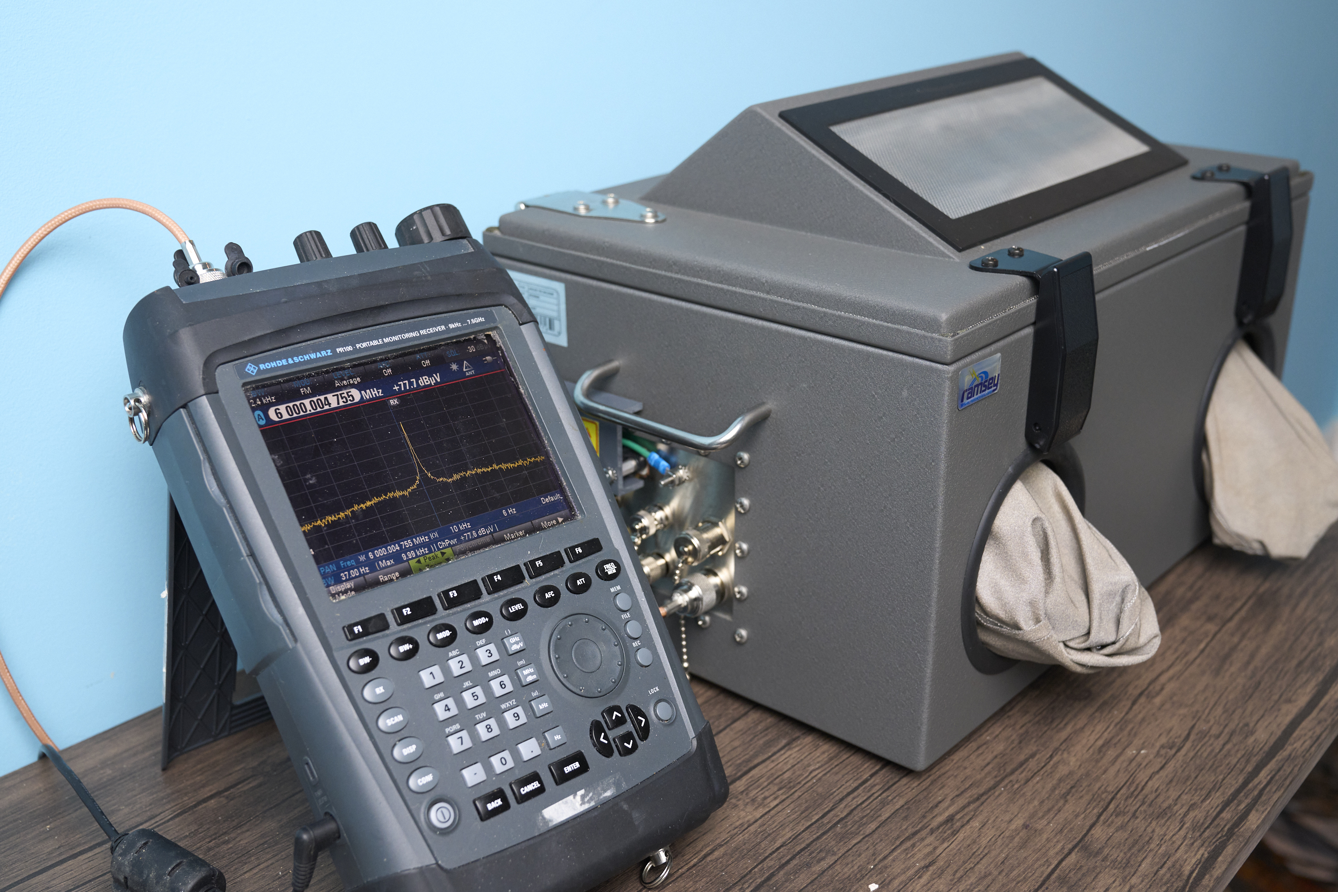 A photo showing a Rohde & Schwarz PR100 portable receiver next to a Ramsey Electronics model STE3000B RF test chamber. The PR100 is a dark grey flat tapered rubber-encased rectangle with lots of buttons and a large screen; the screen displays a fine orange line showing a peak at the center. The STE3000B looks like a cross between a brutalist cash register and an ice box that has turned out its pockets to show that it doesn't have any change. Most of the exterior is metal, in a gunmetal shade of grey that means business.